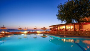 A swimming pool with lounge chairs and umbrellas, a bar area with people, and a view of the sea. - Xenia (Aristotelis)