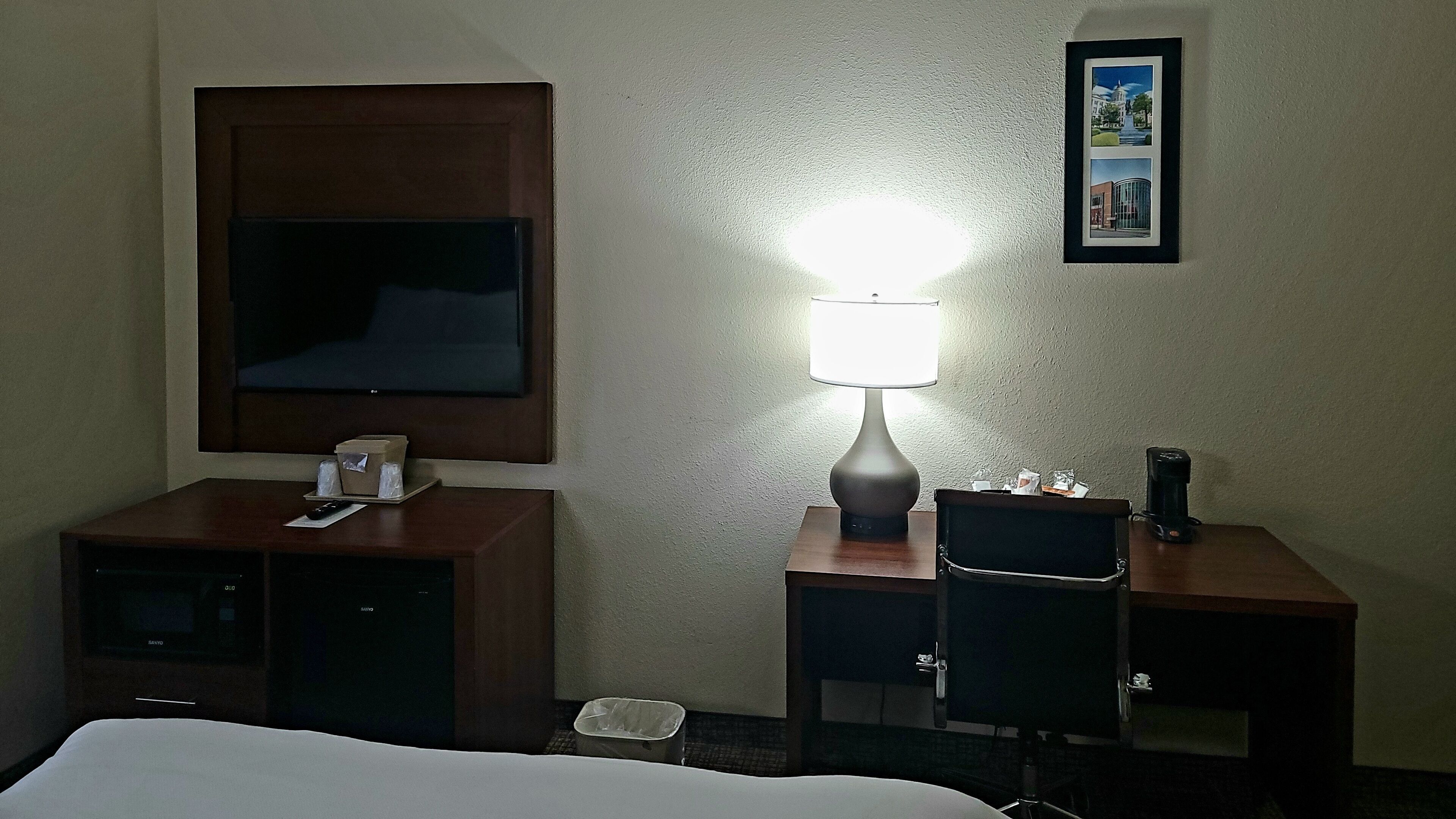 A hotel room with a bed, a desk with a lamp, a chair, a TV, and a framed picture on the wall.