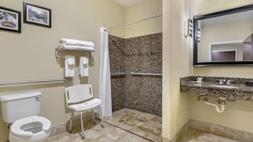 A bathroom with a toilet, a shower with a grab bar, a white chair, and a sink with a mirror.