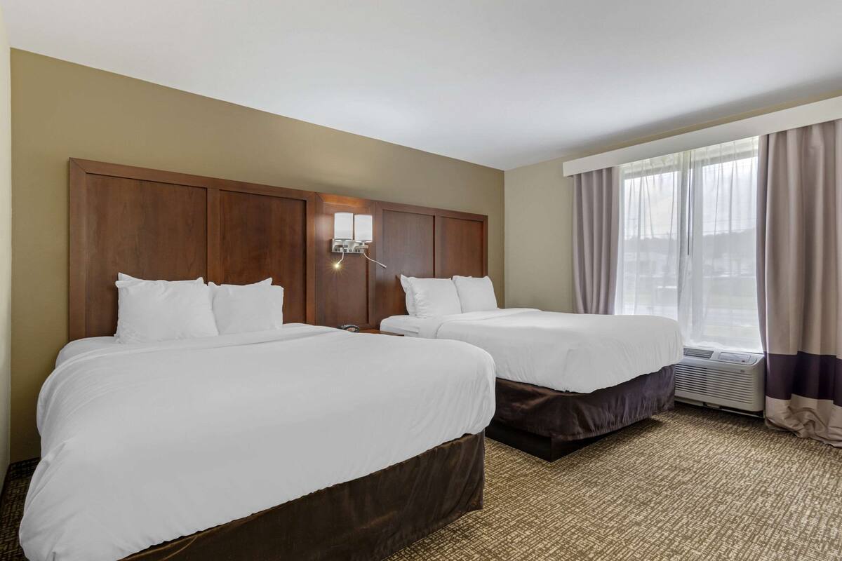 A hotel room with two beds, a wooden headboard, a wall-mounted light, and a large window with curtains.