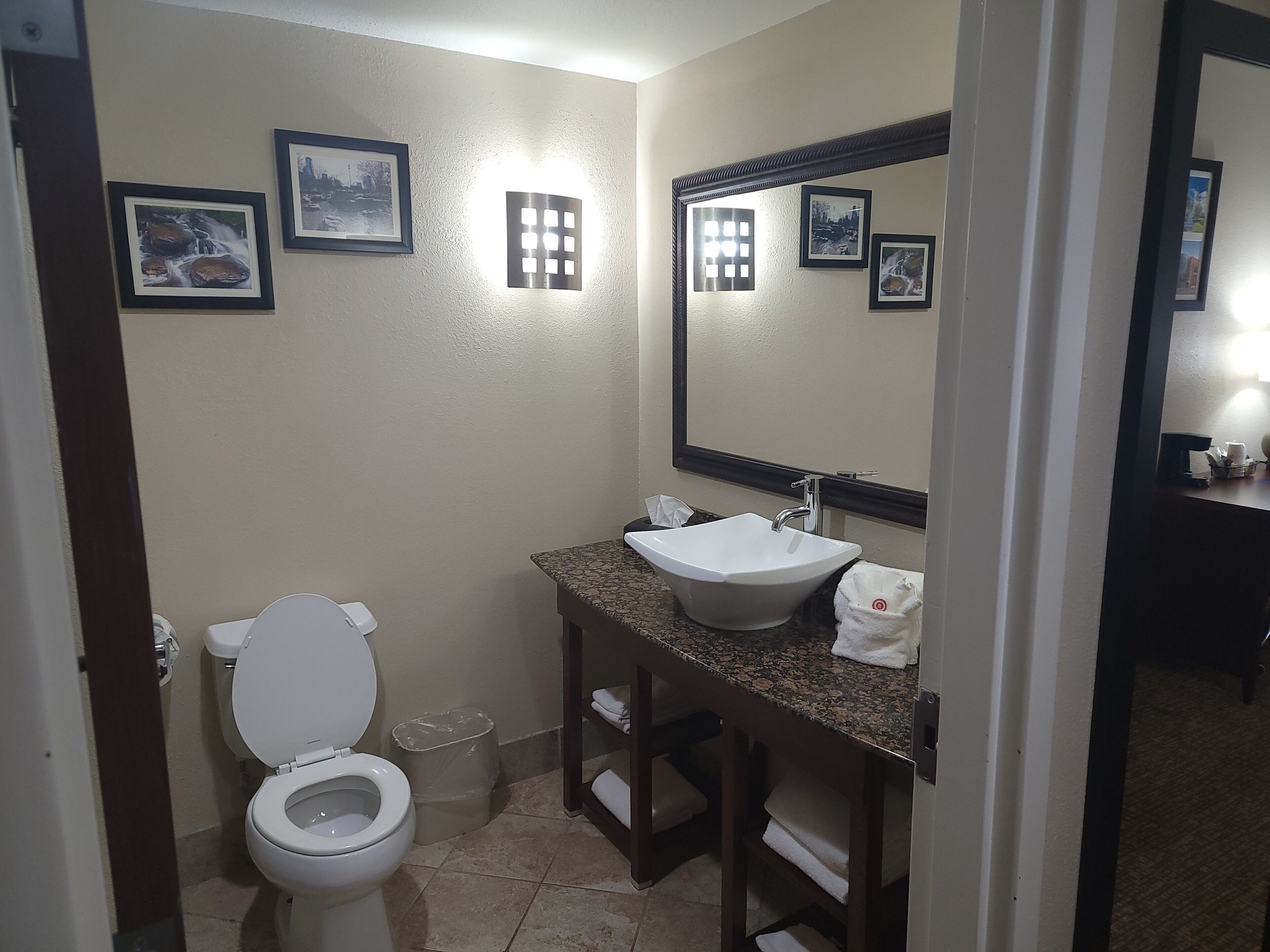A bathroom with a toilet, sink, and mirror. There are framed pictures on the wall and a trash can.