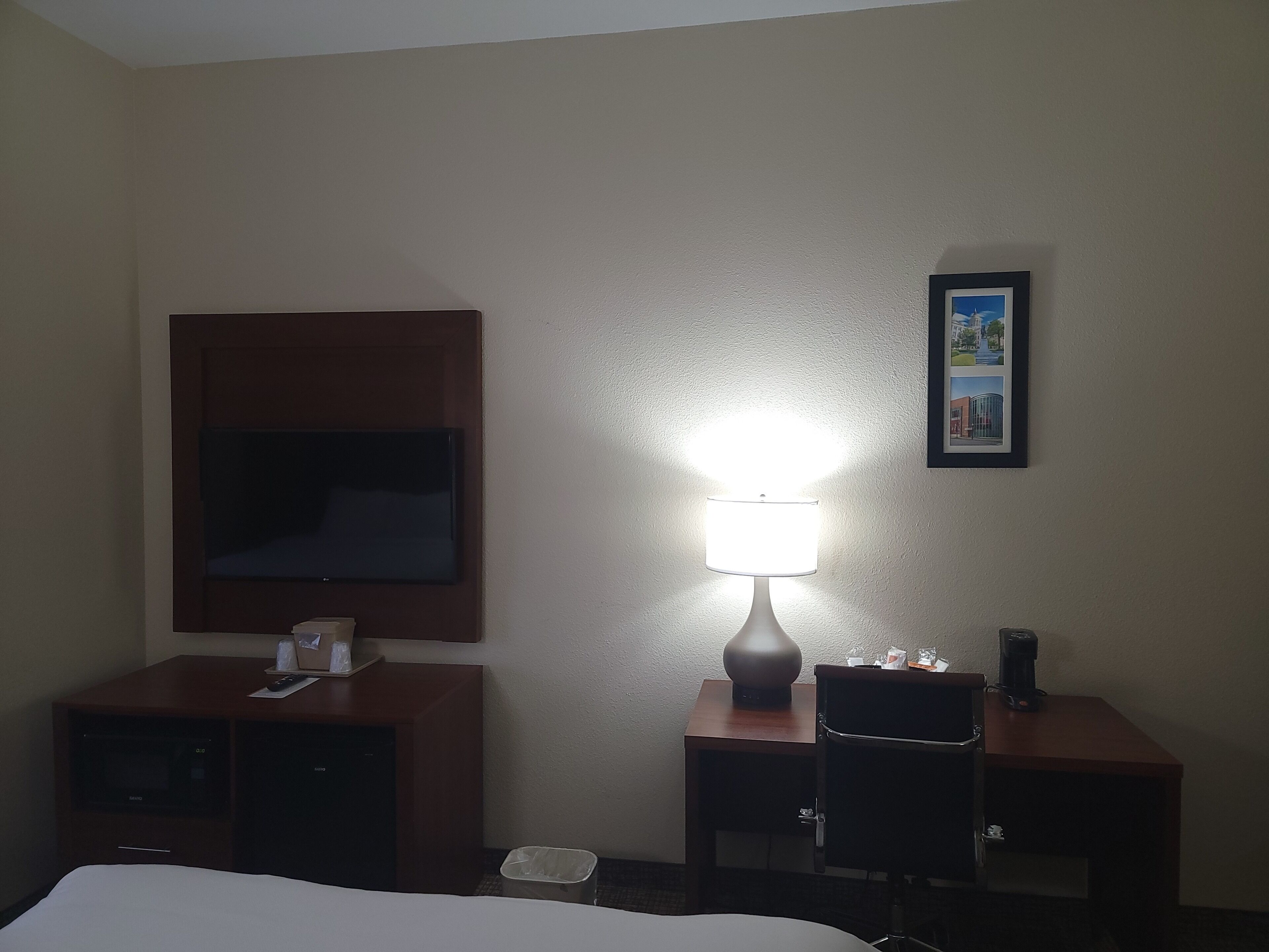 A hotel room with a bed, a TV mounted on the wall, a desk with a lamp, and a small trash bin.