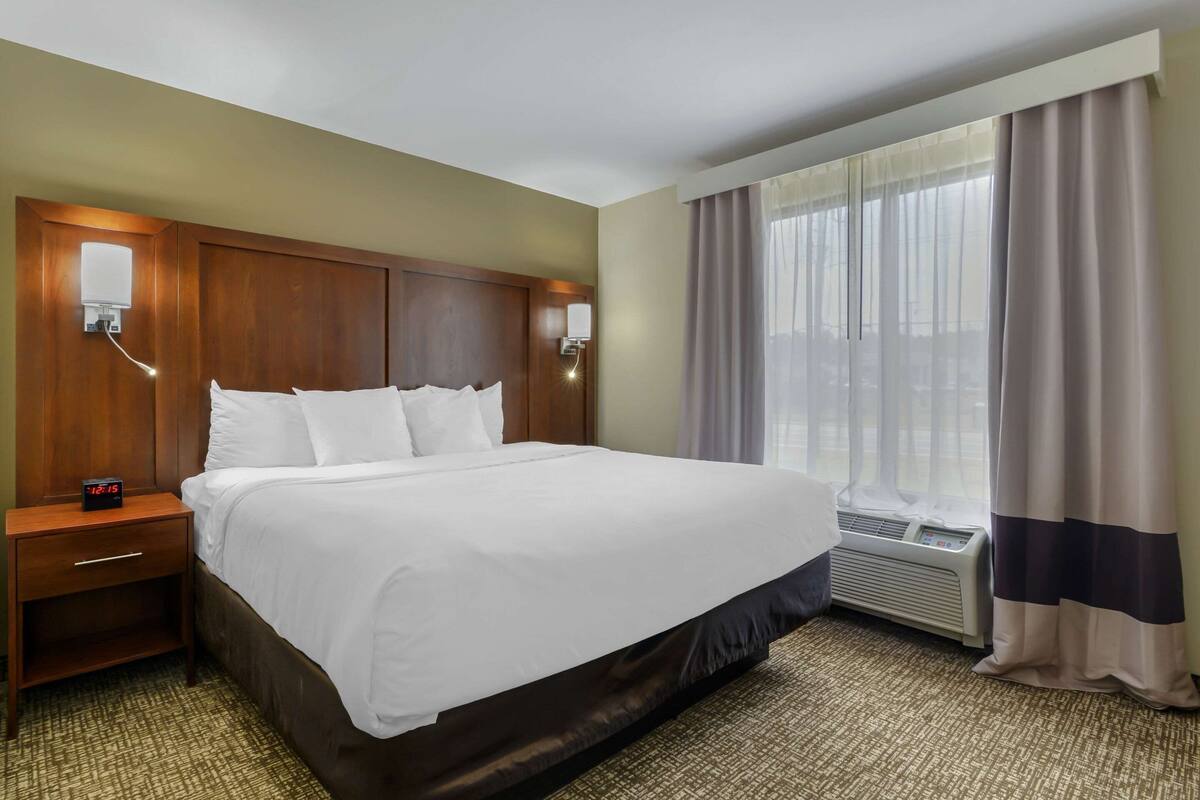 A hotel room with a large bed, a nightstand with a clock, a wall-mounted lamp, and a window with curtains.