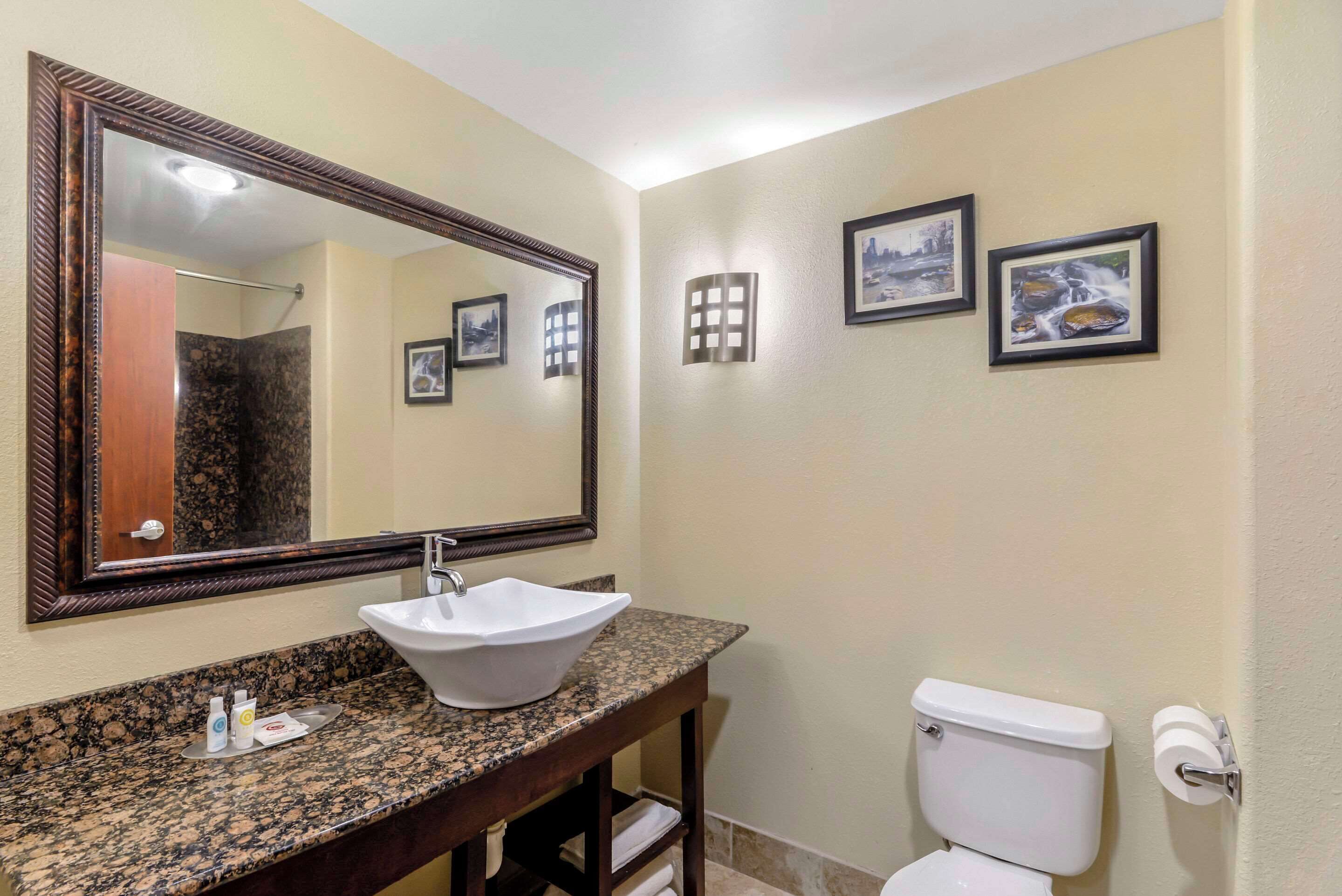 Une salle de bain avec un comptoir en granit, un lavabo blanc, un grand miroir, des toilettes et trois tableaux encadrés accrochés au mur.