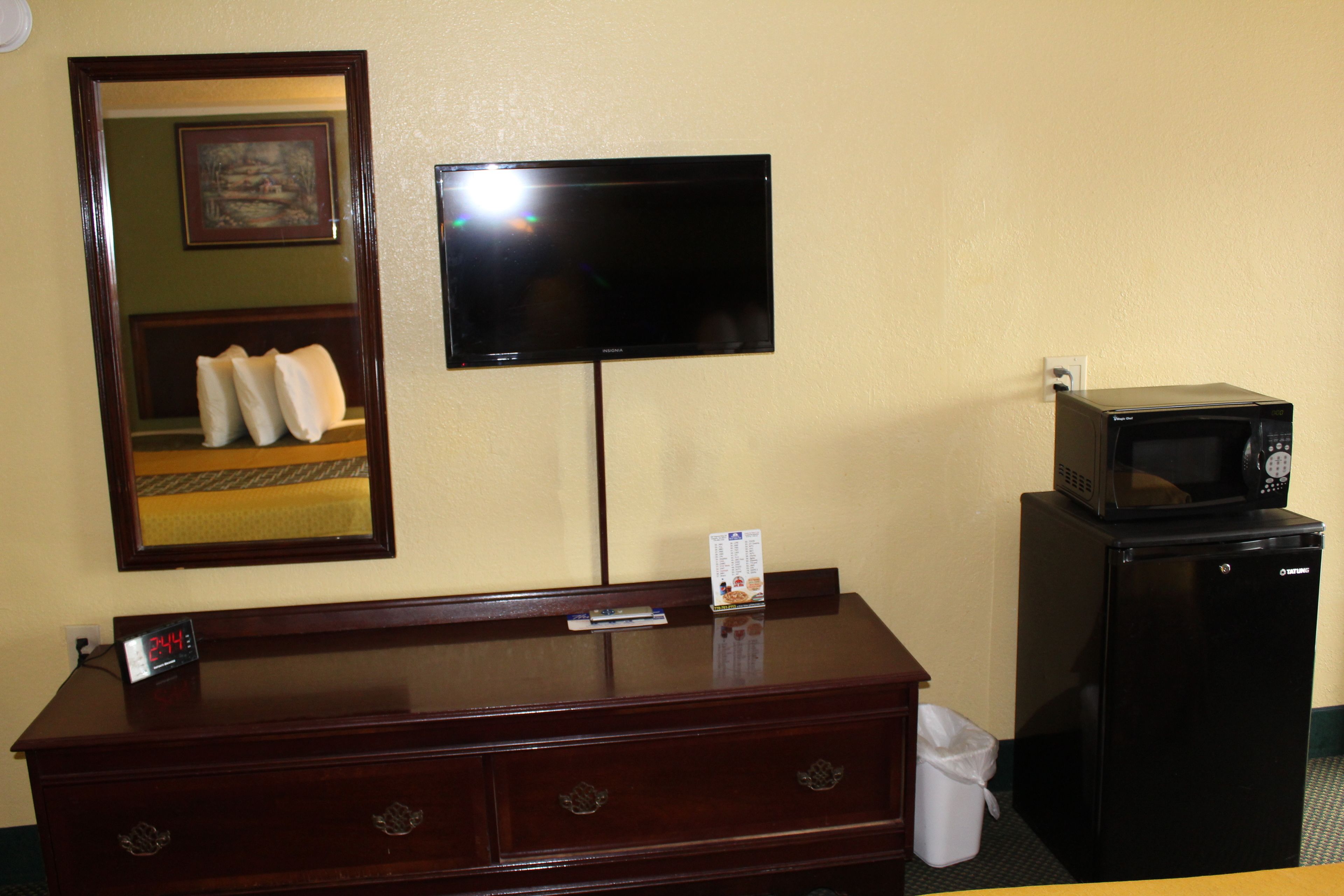 Habitación de hotel con cómoda de madera, televisor de pantalla plana en la pared, microondas sobre una mesita y un cubo de basura.
