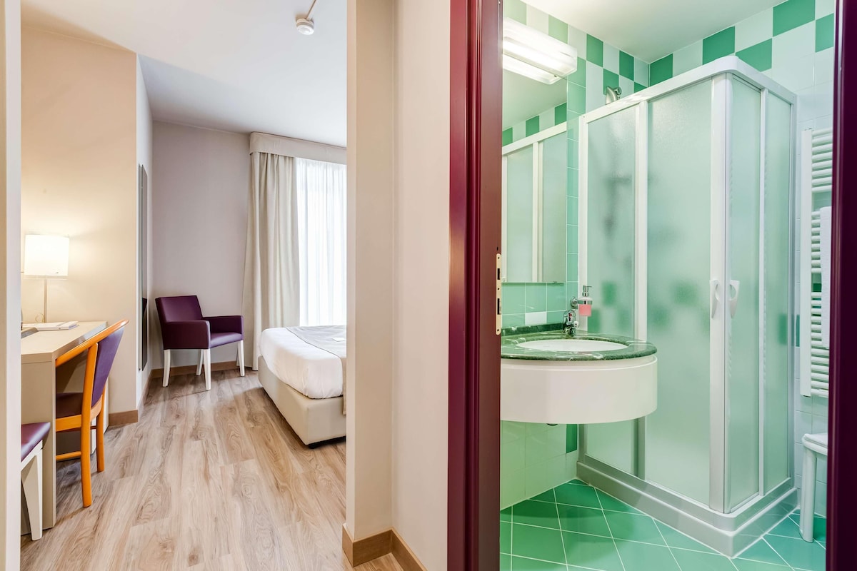 Habitación de hotel con cama, escritorio, silla, ventana con cortinas y baño con lavamanos y ducha.