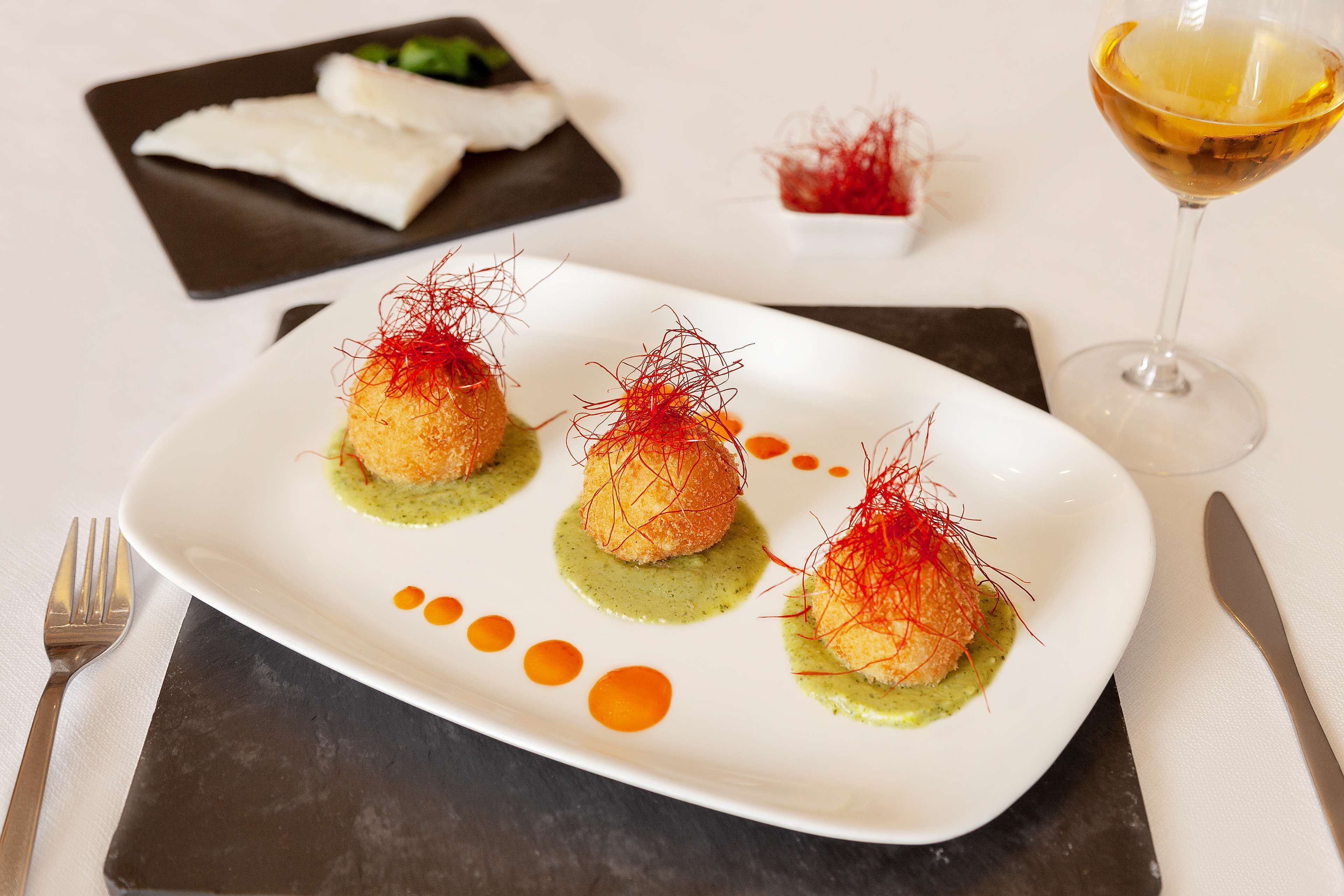 Un plato blanco con tres aperitivos esféricos adornados con hebras rojas, una copa de vino blanco y un pequeño recipiente con guarnición roja sobre una mesa.