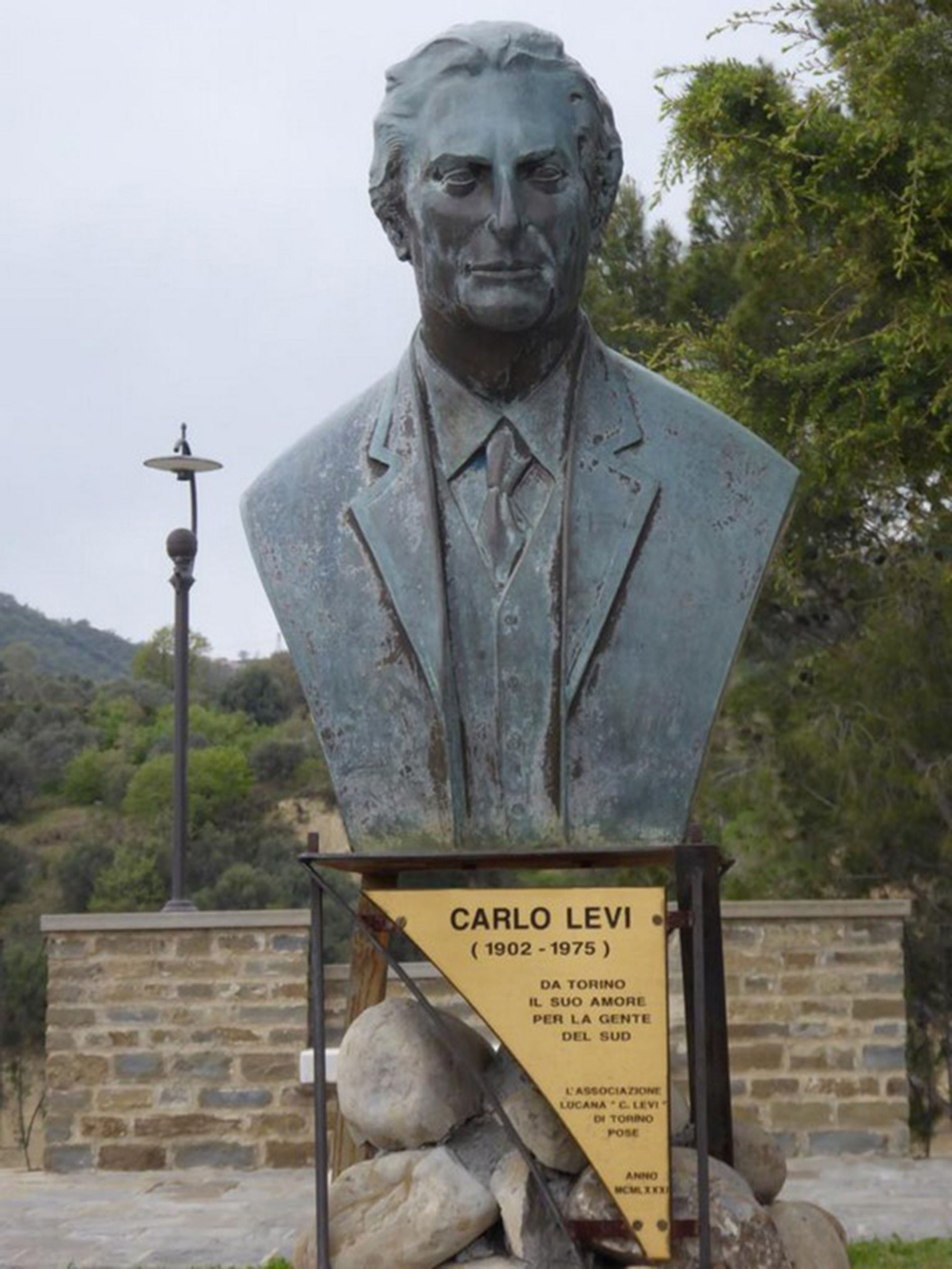 A bust statue of Carlo Levi with a plaque, set against a stone wall and trees.