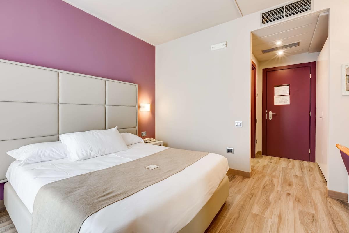 Une chambre d’hôtel avec un grand lit, une tête de lit, une table de chevet, une commode, une lampe fixée au mur, une porte et un plancher en bois.