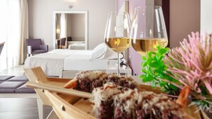 Une chambre d’hôtel moderne avec un lit, une table de chevet en bois sur laquelle se trouve un aliment, et deux verres de vin blanc.