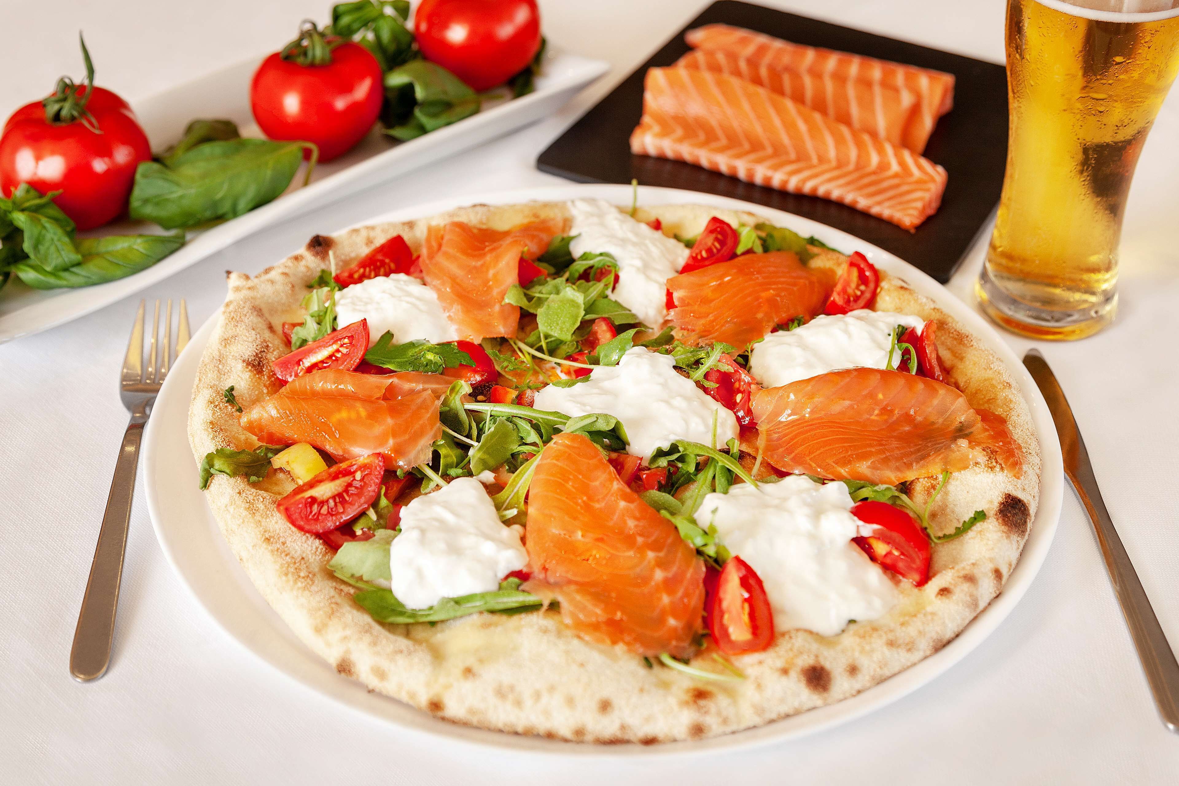 A plate of food with salmon, tomatoes, and greens, a glass of beer, and a fork and knife on a table.