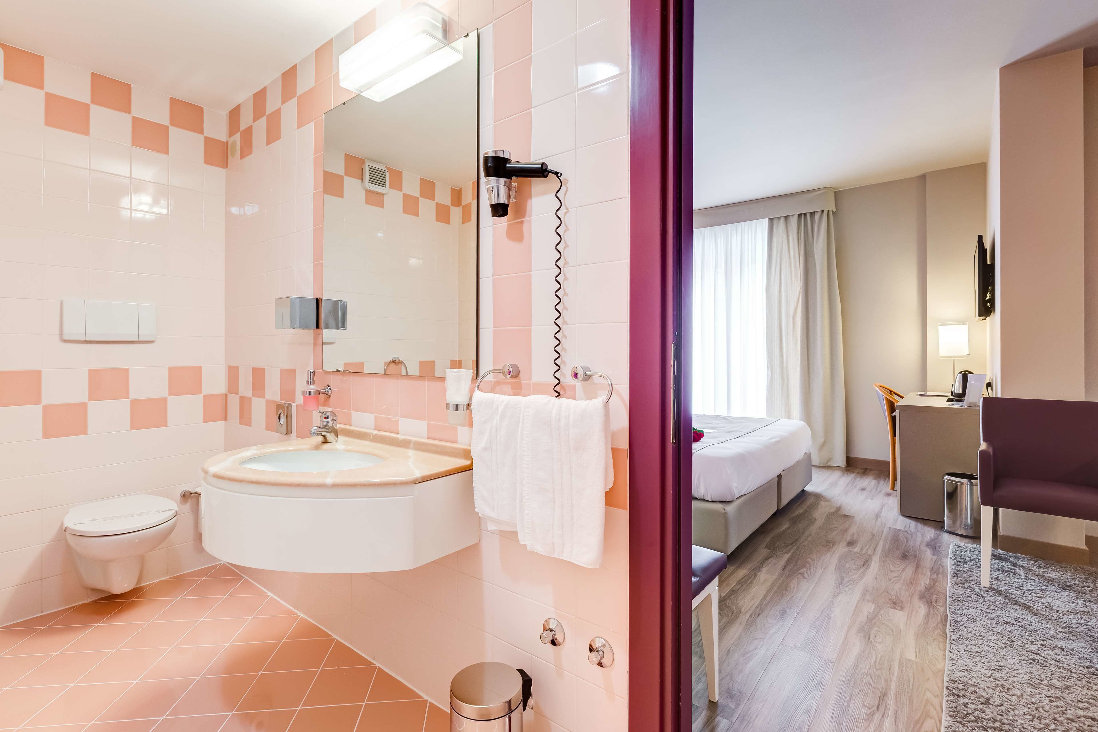 A modern bathroom with a sink, toilet, and shower. There is a bed and a desk in the adjacent room.