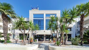 Un hôtel moderne, reconnaissable à son grand panneau lumineux indiquant « HÔTEL », doté d’une entrée couverte et d’un espace extérieur bien entretenu, agrémenté de palmiers et de plantes en pot.