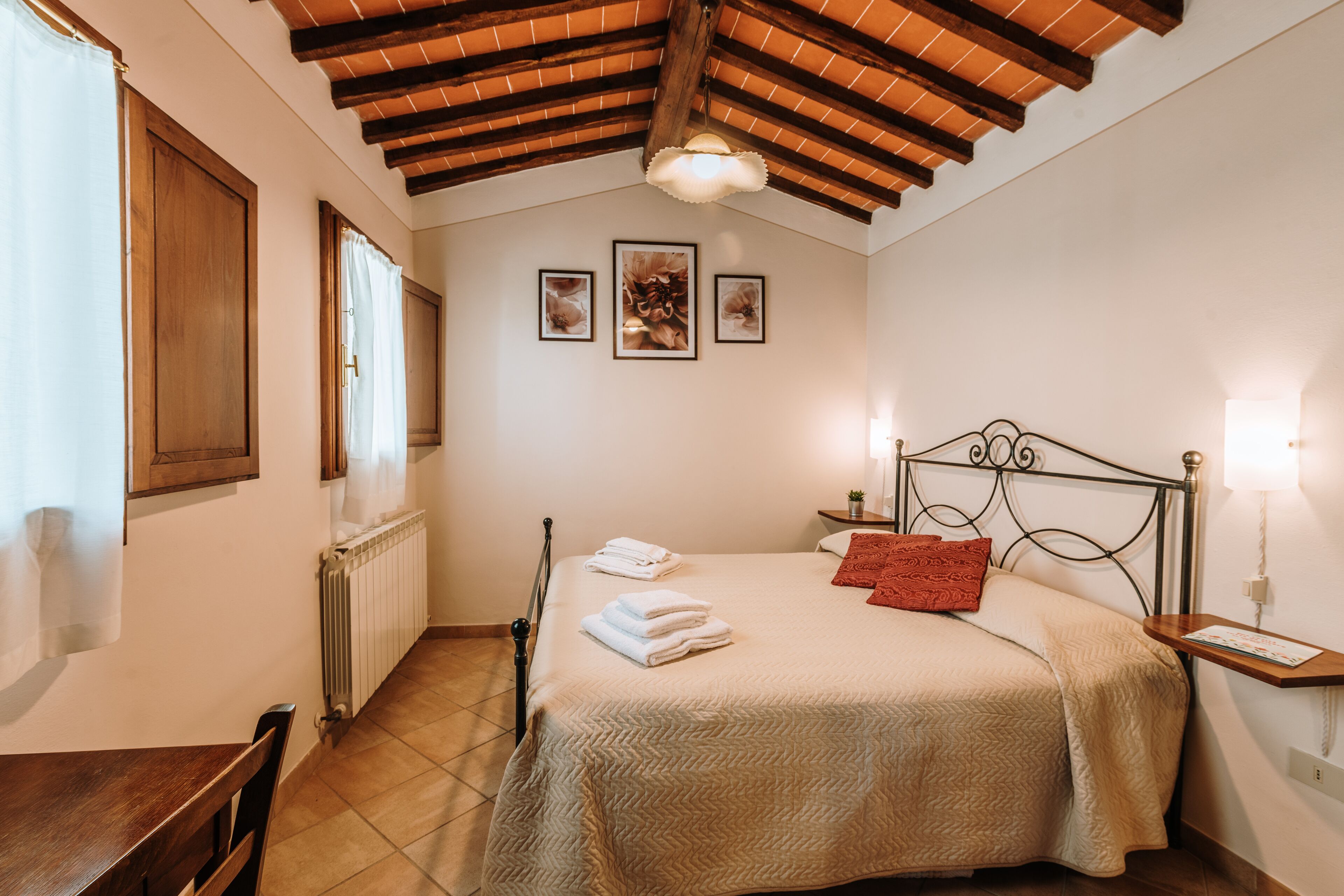 Een slaapkamer met een houten plafond, een bed met wit en rode beddengoed, een nachtkastje met een lamp en een raam met gordijnen.