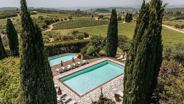 A pool area with two rectangular pools, surrounded by lounge chairs and umbrellas, set against a backdrop of vineyards and tall cypress trees. - Villa di Sotto (Castelnuovo Berardenga)