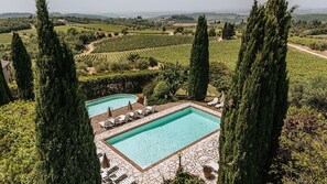 A pool area with two rectangular pools, surrounded by lounge chairs and umbrellas, set against a backdrop of vineyards and tall cypress trees. - Villa di Sotto (Castelnuovo Berardenga)