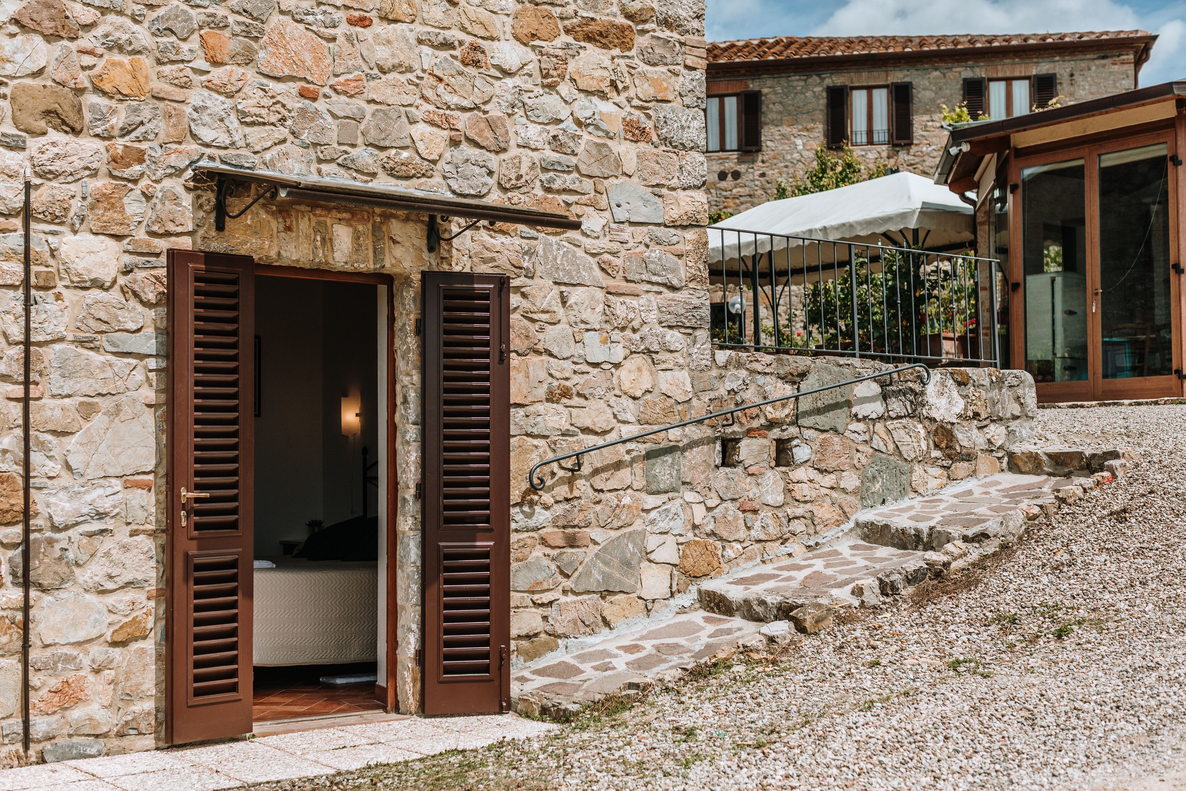 A stone building with a wooden door and shutters, a staircase leading to a balcony with a white umbrella, and a second building in the background.