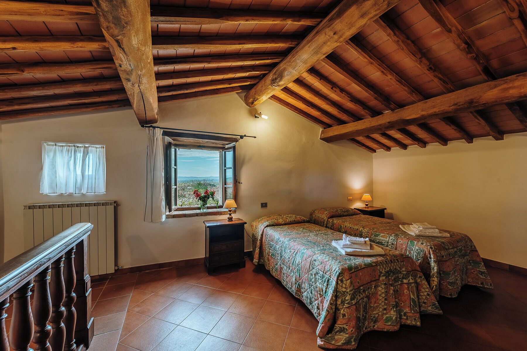 A rustic room with a sloped ceiling, two beds with patterned bedding, a window with a view, and a radiator.
