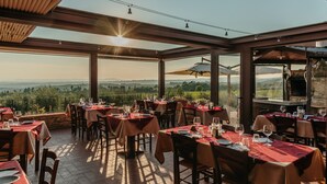 A restaurant with tables set for dining, large windows offering a view of the landscape, and a wooden ceiling structure. - Villa di Sotto (Castelnuovo Berardenga)
