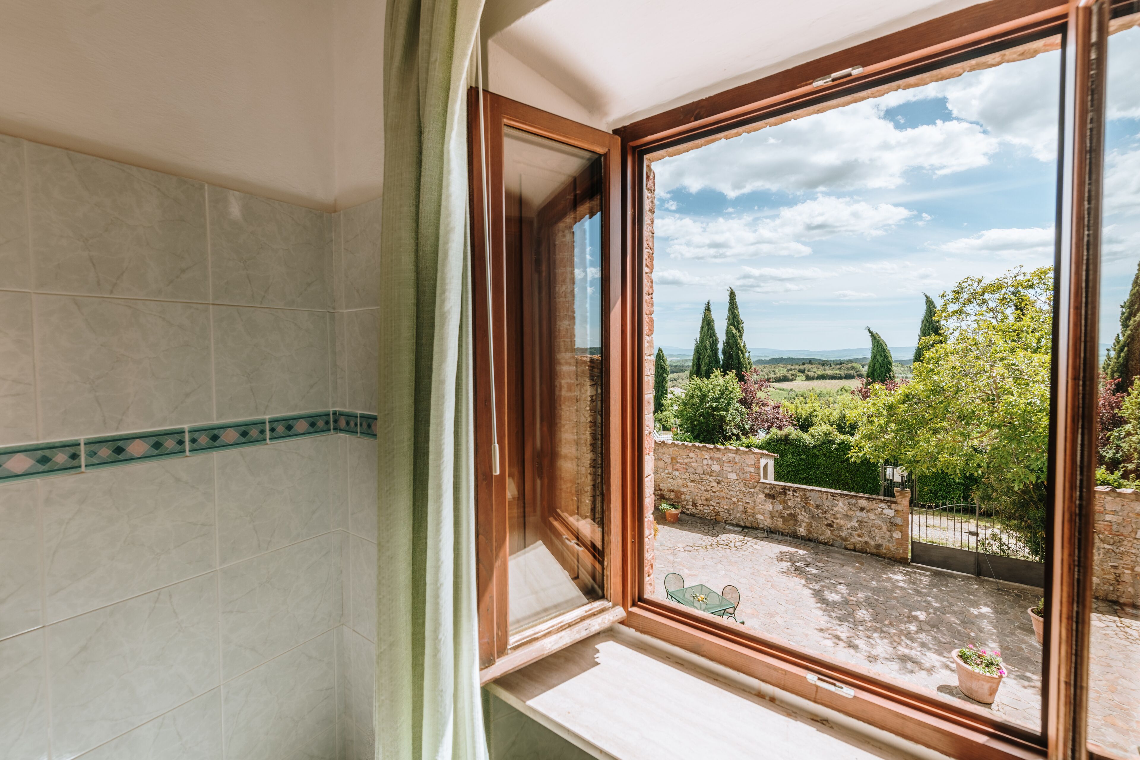 Ein Badezimmer mit großem Fenster, gefliesten Wänden und Blick auf einen Innenhof mit Grünflächen und einer Steinmauer.