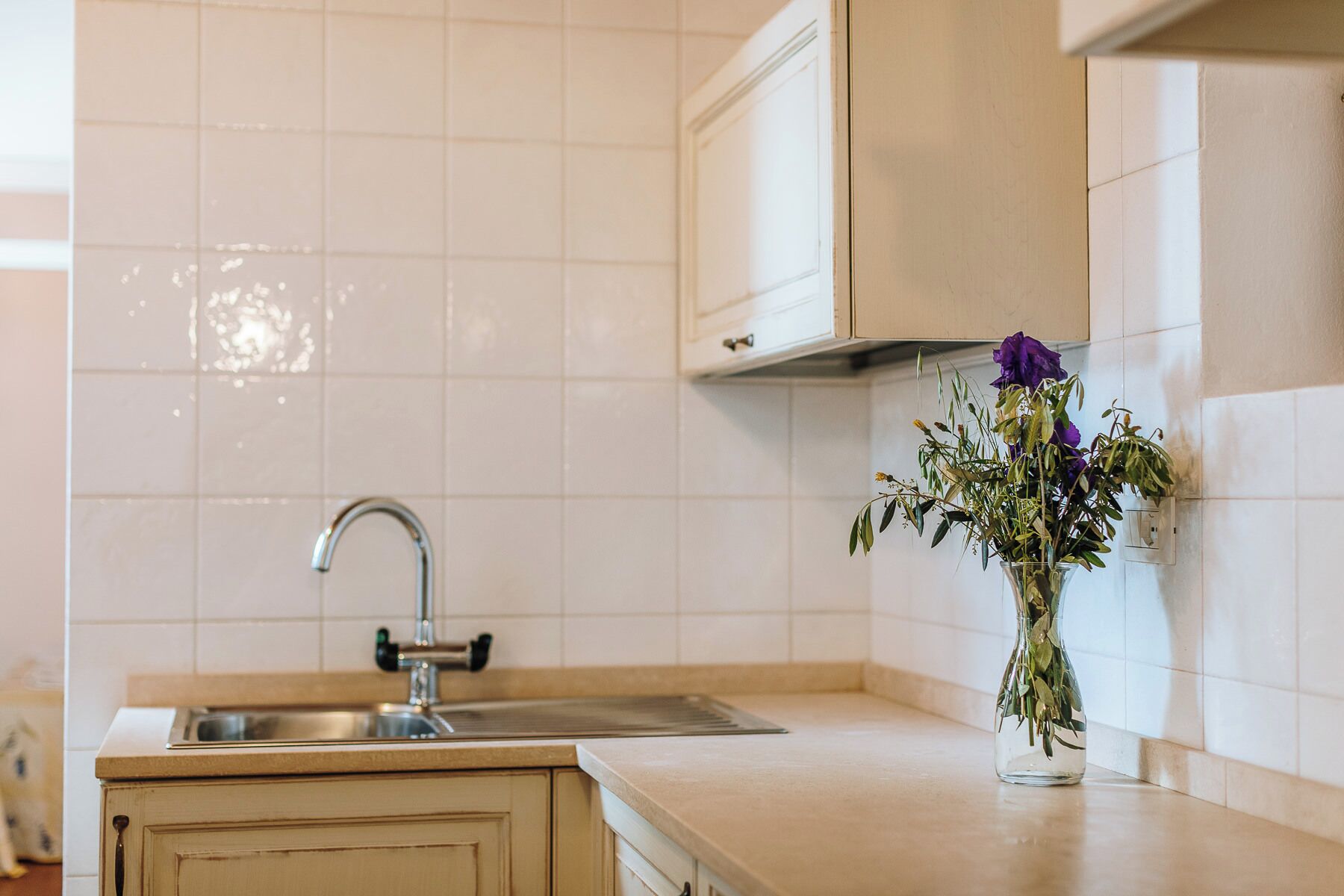 Et køkken med hvide fliser, en stålvask og en vase med blomster på bordet.