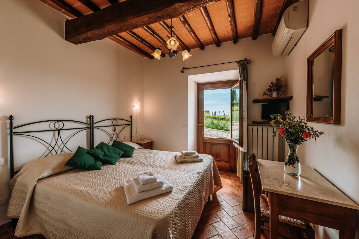 Ein Schlafzimmer mit einem Bett, einer Holzbalkendecke, einem Fenster, einem Tisch mit einer Vase und einem Spiegel.