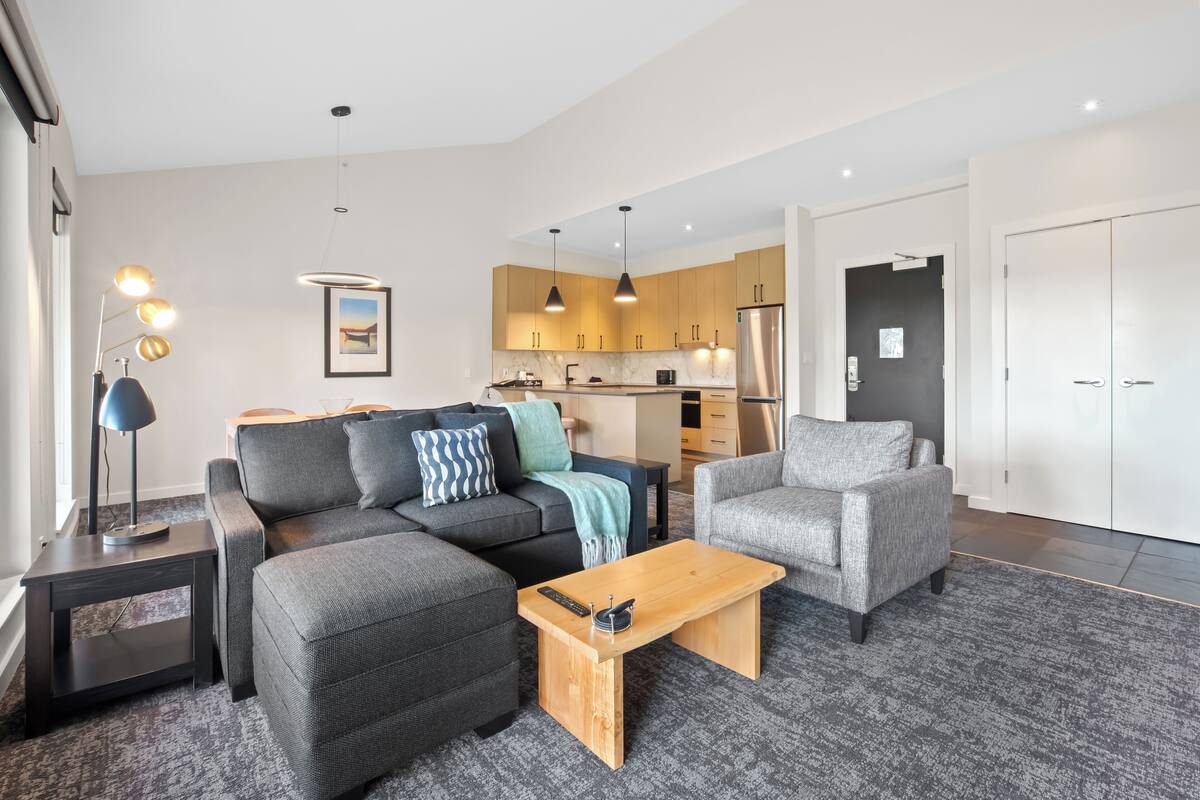 Een moderne woonkamer met een bank, fauteuil en salontafel, een keuken op de achtergrond, en een deur die naar een andere kamer leidt.