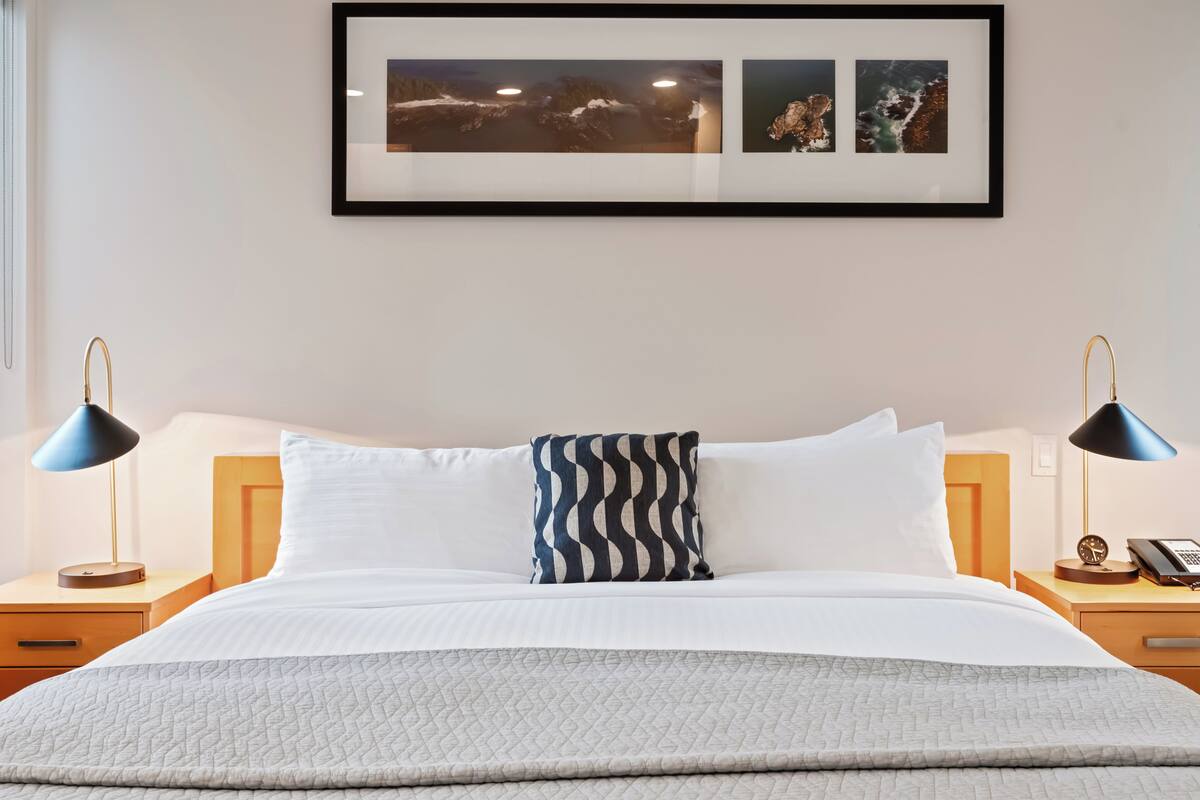 Een net opgemaakt bed met een gewatteerd dekbed, twee bedlampjes, een nachtkastje met een telefoon en ingelijste foto's aan de muur.
