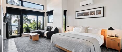 A modern bedroom with a large bed, a sofa, a small table, and a nightstand. There is a window with a view of the outdoors.