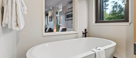 Un baño moderno con una bañera independiente, un espejo grande y una ventana con vistas a árboles.