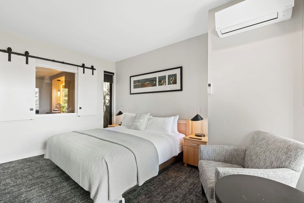 Habitación de hotel con cama, mesitas de noche, una silla, una pequeña mesa, equipo de aire acondicionado y un cuadro en la pared.