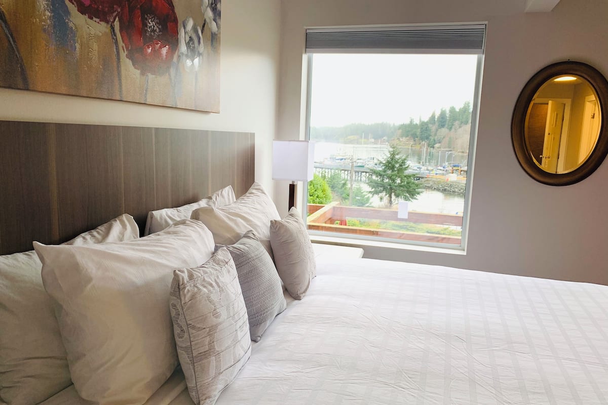 Une chambre d’hôtel avec un grand lit, une tête de lit, une fenêtre donnant sur un quai et des arbres, et un miroir rond accroché au mur.