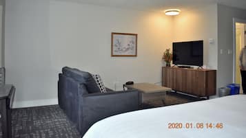 A modern hotel room with a grey sofa, a small wooden table, a flat-screen TV on a wooden stand, a framed picture on the wall, and a bed with a white comforter.