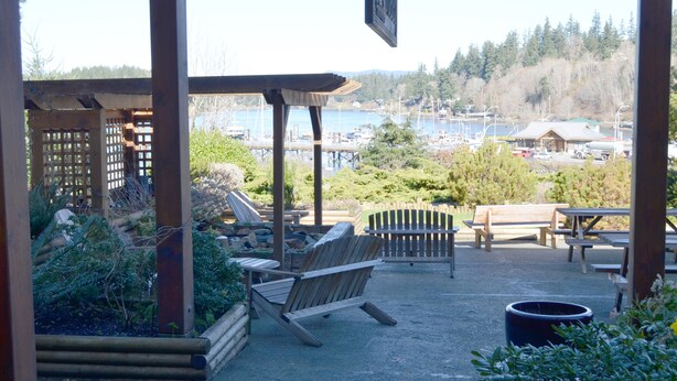 Un espace extérieur aménagé avec des bancs et des chaises en bois, une table de pique-nique couverte, et une vue sur une marina avec des bateaux, ainsi qu’une colline boisée en arrière-plan.