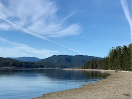 Un lago tranquilo con una playa de guijarros y montañas boscosas al fondo.