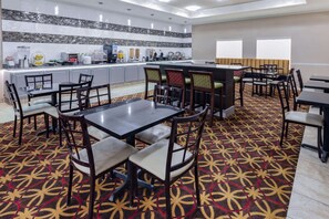 Un comedor de hotel con alfombra estampada, varias mesas y sillas, una zona de cocina con electrodomésticos y un ventanal que proporciona luz natural.