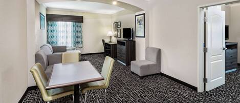 A hotel room with a dining area, a sofa, a TV, and a door leading to another room.