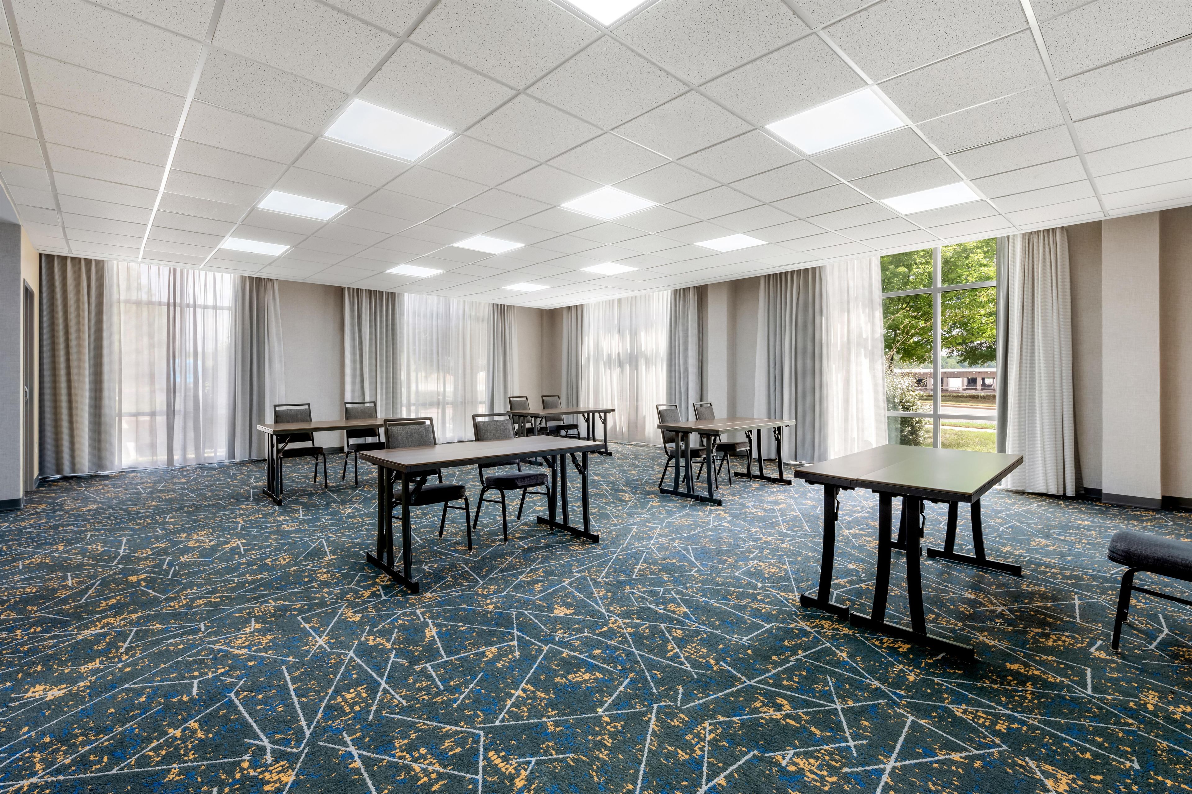 A conference room with a high ceiling, rectangular tables, and chairs, large windows with curtains, and a patterned carpet.