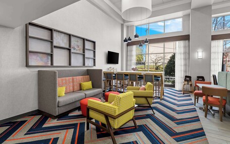A modern hotel lobby with a gray sofa, yellow and red chairs, a dining area with tables and chairs, and a large window offering a view of the outdoors.