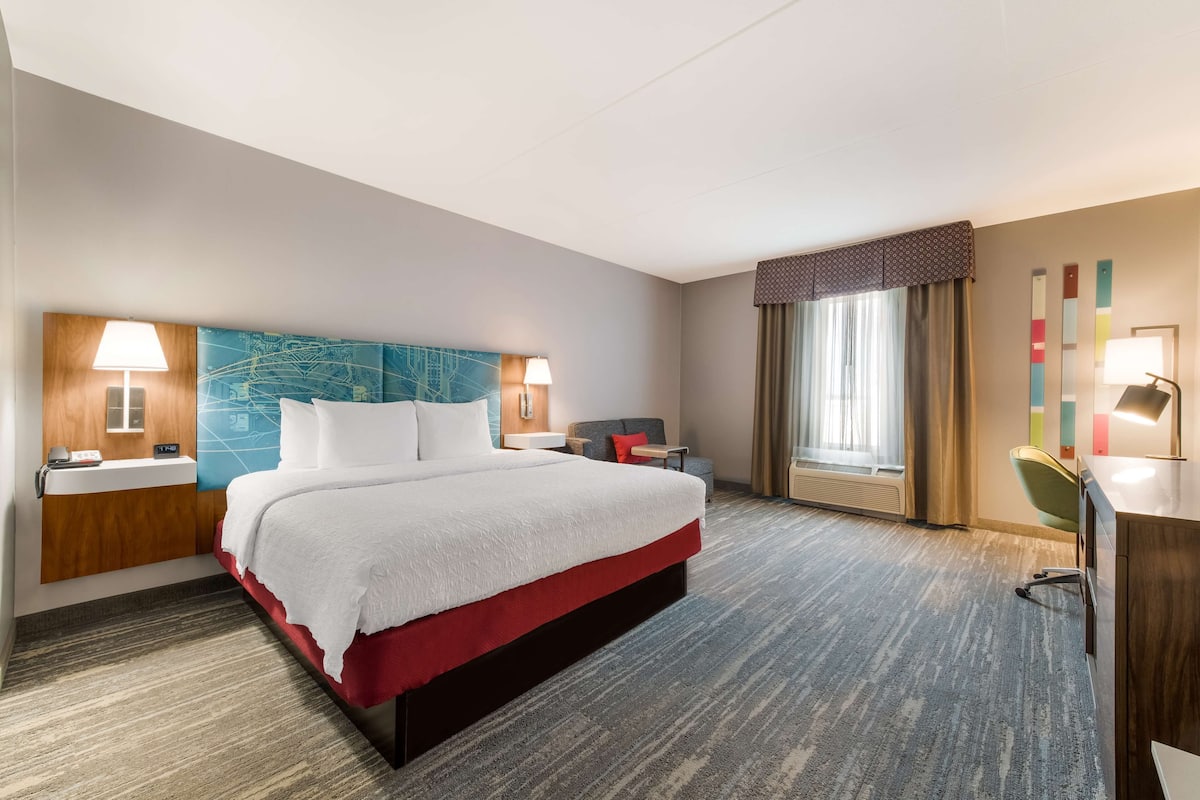 A hotel room with a bed, bedside tables, a desk, a chair, a window with curtains, and a wall with a colorful abstract design.