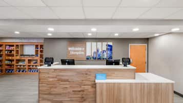 A hotel reception area with a wooden reception desk, two computer monitors, a 'welcome to Hampton' sign, and a 'Hilton Honors' sign.
