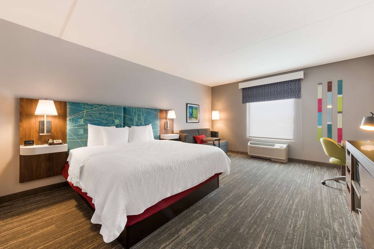 A hotel room with a large bed, bedside tables, a desk with a chair, a window with blinds, and a wall with a colorful abstract design.