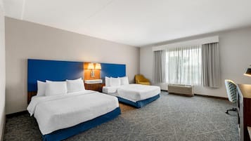 A hotel room with two beds, a desk, a chair, a window with curtains, and a wall-mounted heater.