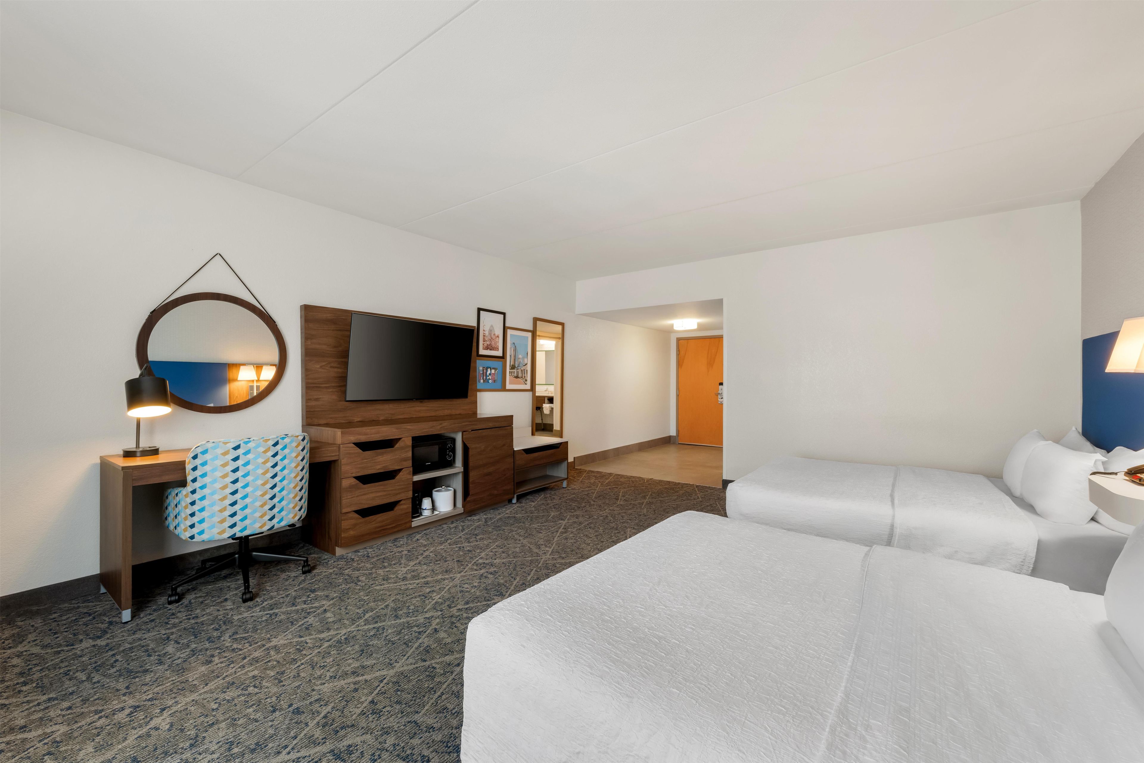 A hotel room with a bed, a desk with a chair, a TV, and a bathroom visible in the background.