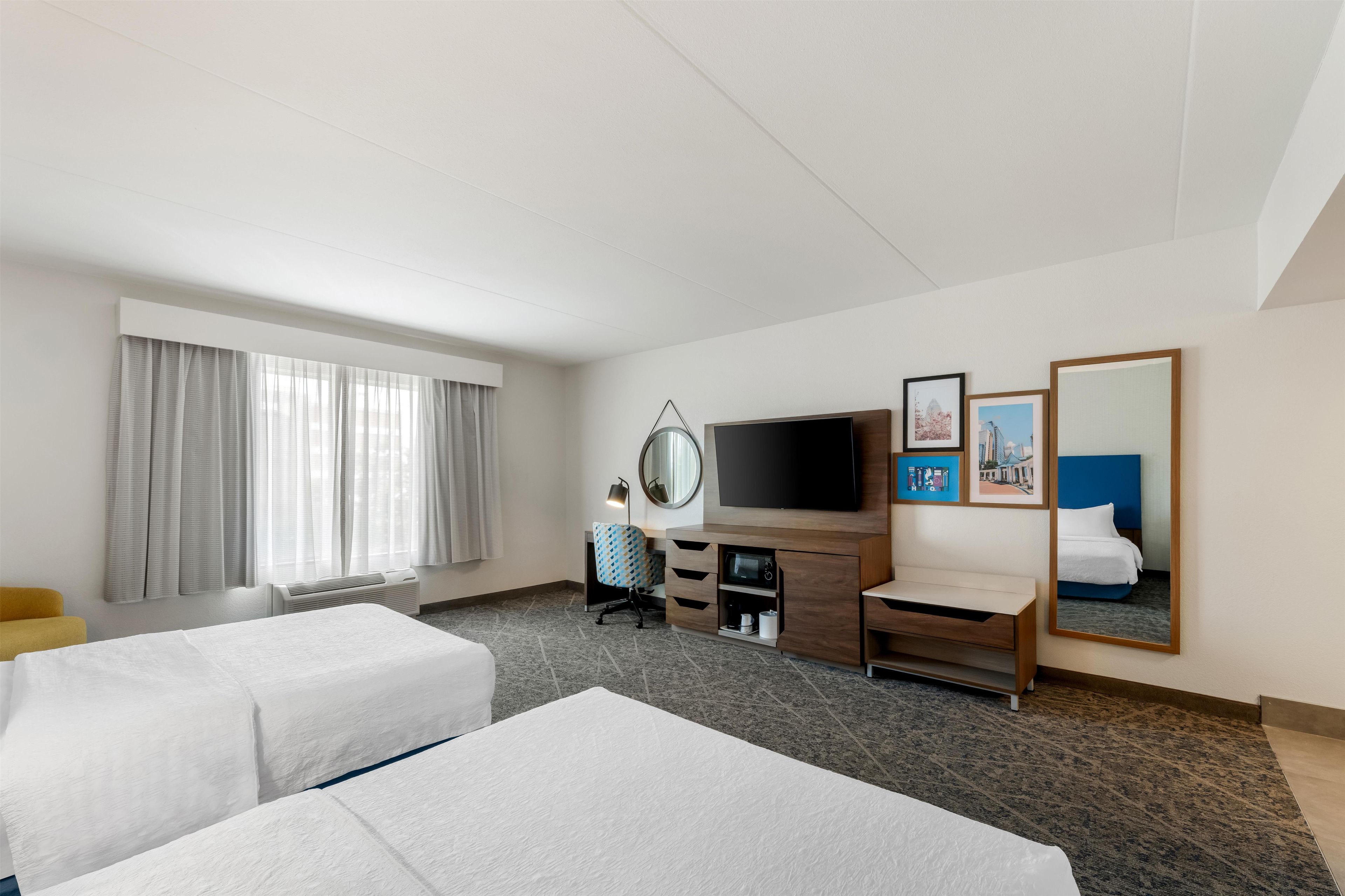 Habitación de hotel con dos camas, televisión, escritorio con silla, espejo y obras de arte en la pared.