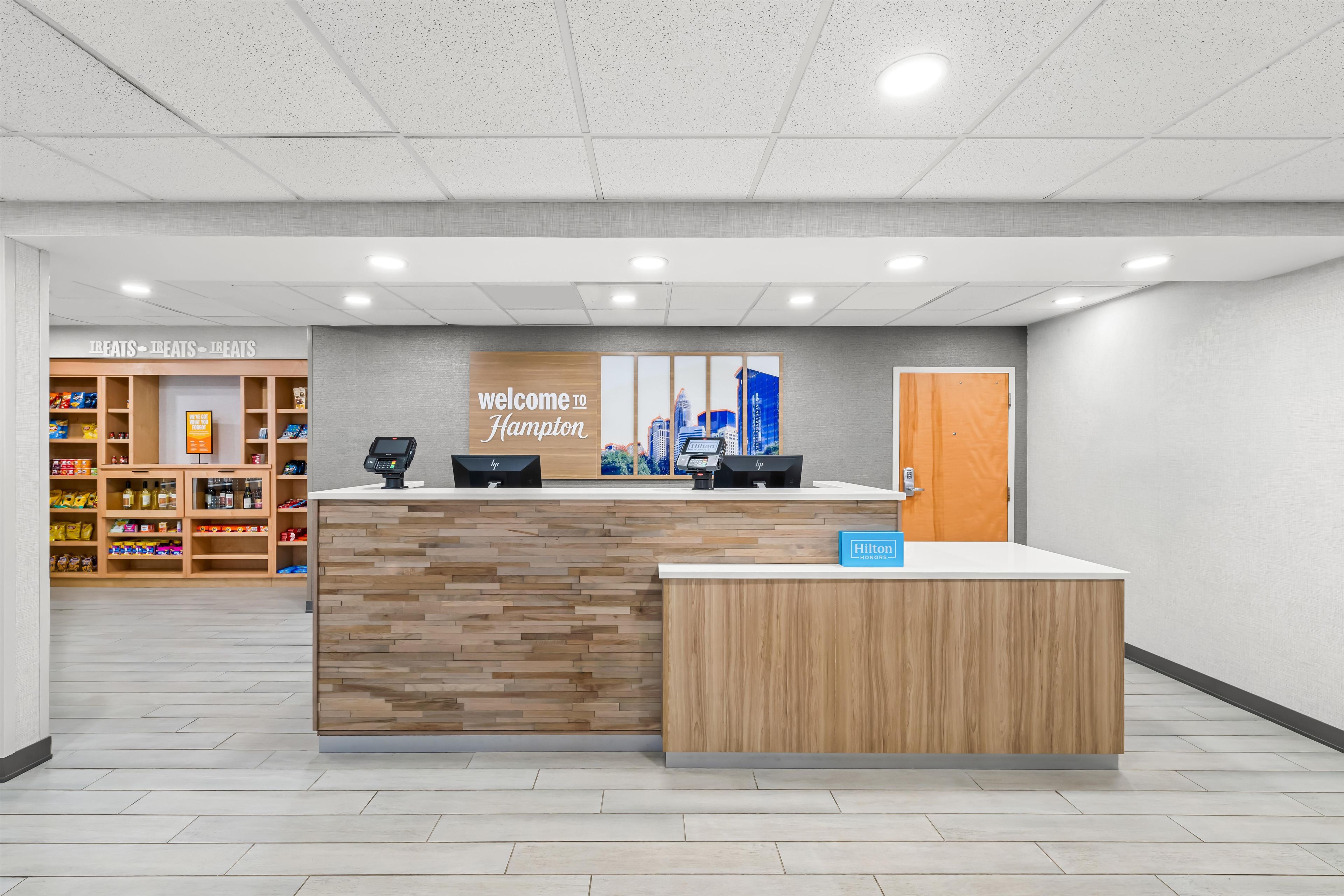 A hotel reception area with a wooden reception desk, a 'Welcome to Hampton' sign, a 'Hilton Honors' sign, and a snack bar in the background.