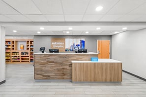 A hotel reception area with a wooden reception desk, a 'Welcome to Hampton' sign, a 'Hilton Honors' sign, and a snack bar in the background. - Hampton Inn & Suites Charlotte Airport (Charlotte)