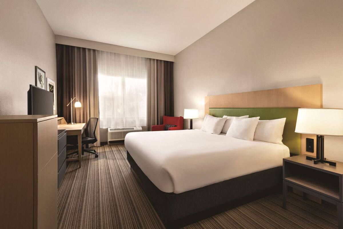 A hotel room with a large bed, a desk with a chair, a red chair, a lamp, and a television on a cabinet.