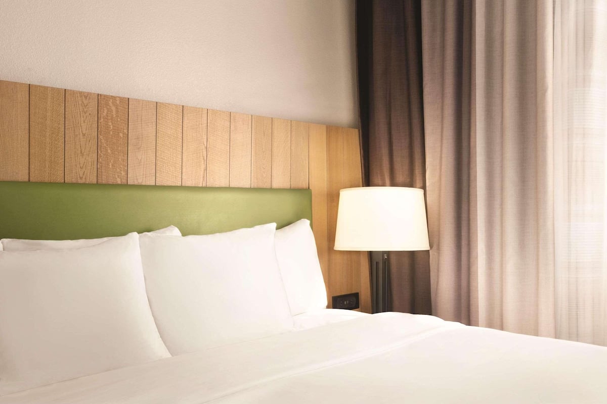 A hotel room with a neatly made bed, a wooden headboard, a green accent wall, and a lamp on the nightstand.