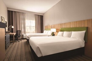 Ein Hotelzimmer mit zwei Betten, einem Schreibtisch, einem Stuhl, einer Lampe und einem großen Fenster mit Vorhängen.